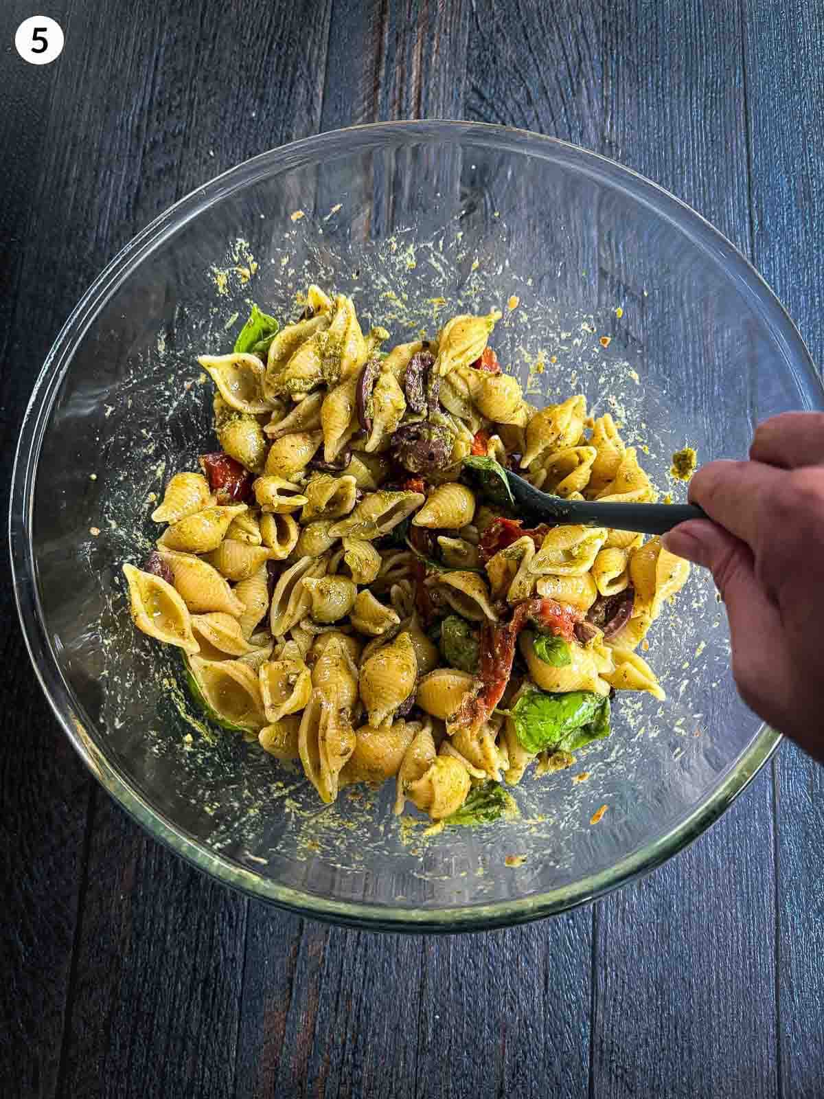stirring ingredients for Semi-dried Tomato Pesto Pasta Salad in a clear bowl with a spatula