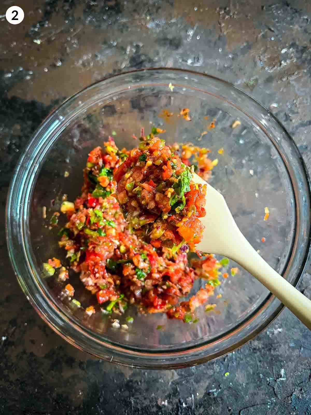lifting a serve of chopped ingredients for ezme salad in a clear bowl, with a silicon spatula